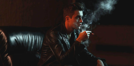 Man wearing black leather jacket smoking cigarette with visible smoke in dimly lit room
