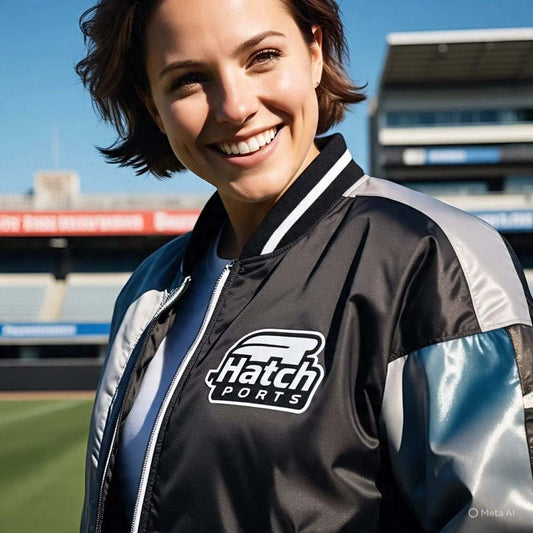 Smiling woman wearing black and silver varsity letterman jacket with Hatch Sports logo at sports stadium
