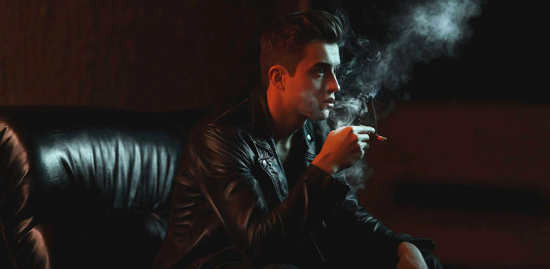 Man wearing black leather jacket smoking cigarette with visible smoke in dimly lit room