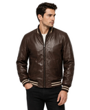 A man wearing a dark brown leather bomber jacket with beige and white striped ribbed trim, paired with black pants, against a light gray studio background.
